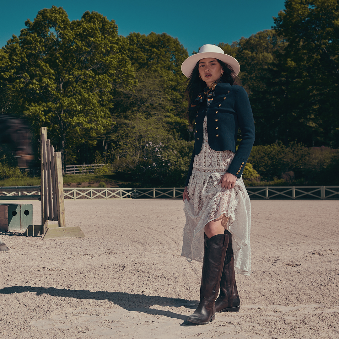 A woman stands in a sandy outdoor arena wearing a white wide-brim hat, dark navy cropped jacket with gold buttons, a white lace dress, and dark brown knee-high boots. Trees and fencing are visible in the background.