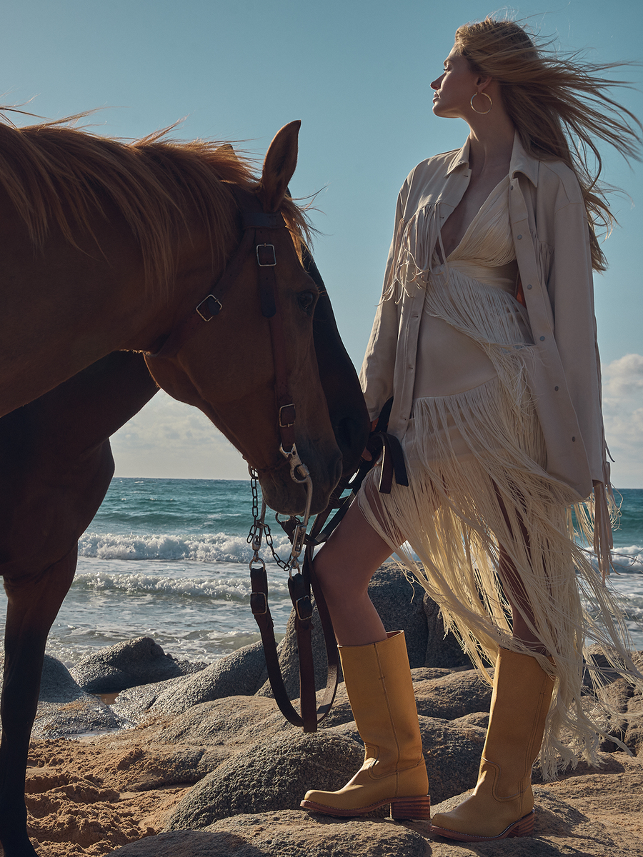 A woman in a cream fringe jacket and matching boots stands on a rocky beach next to a brown horse, with the ocean and blue sky in the background.