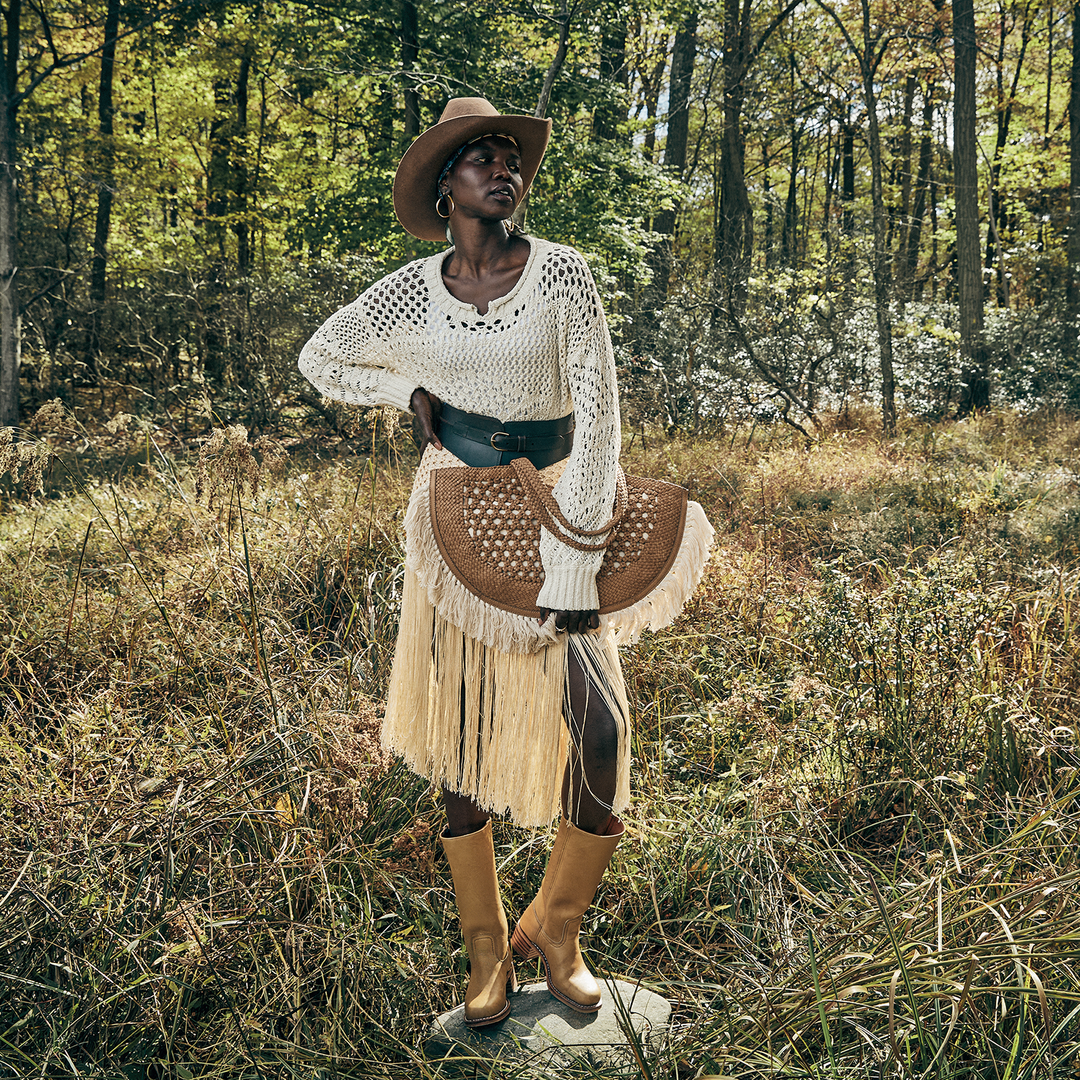 A model stands in a grassy forest clearing wearing a cream crochet sweater, a brown fringe skirt, a wide black belt, tan boots, and a brown cowboy hat, while holding a matching brown woven hat.