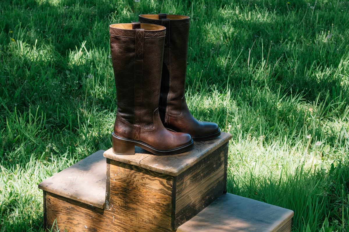 A pair of dark brown leather knee-high riding boots displayed on a rustic wooden step outdoors on a grassy lawn.
