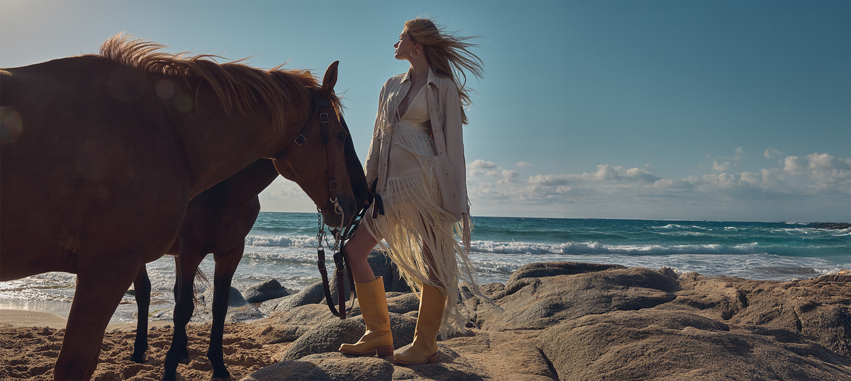 A woman in a white fringed coat and tan boots stands on a rocky beach next to a brown horse, with the ocean and blue sky in the background.