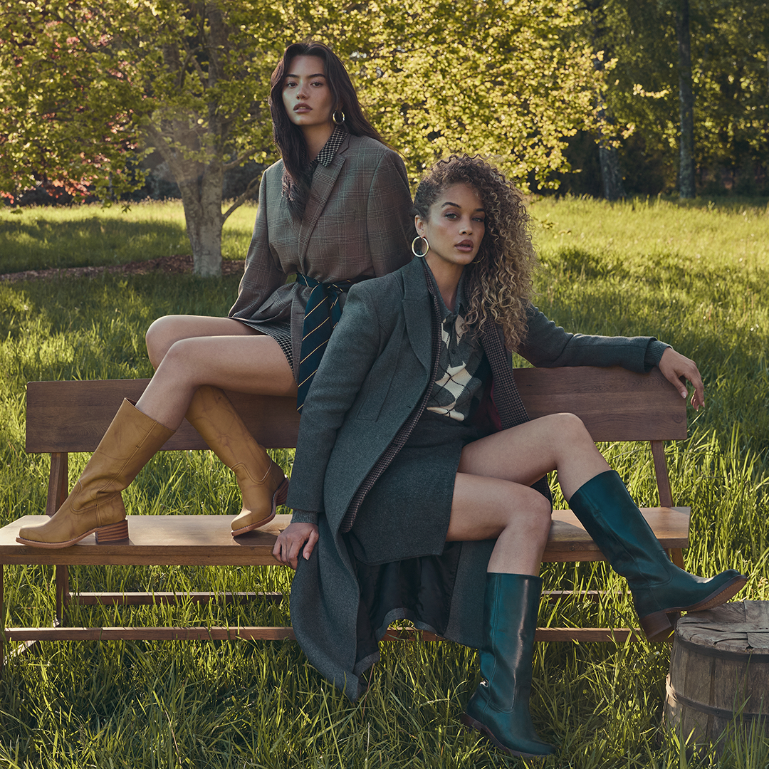 Two models posing on a wooden bench in a grassy park setting, wearing coordinated dark outerwear, patterned tops, and tall boots.