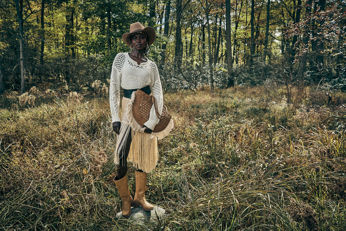 A person stands in a grassy woodland clearing wearing a cream knit long-sleeve top, a beige fringe skirt, tan cowboy boots, and a brown hat while holding a woven brown hat. Trees and brush fill the background.