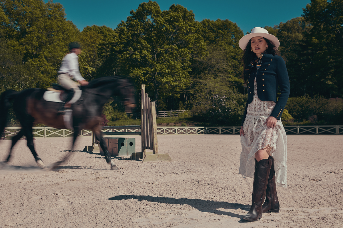 A woman in a white cowboy hat, dark blue jacket, white lace skirt, and brown riding boots stands in a sandy equestrian arena. A rider on a dark horse is blurred in the background near a jump, with trees and blue sky behind her.